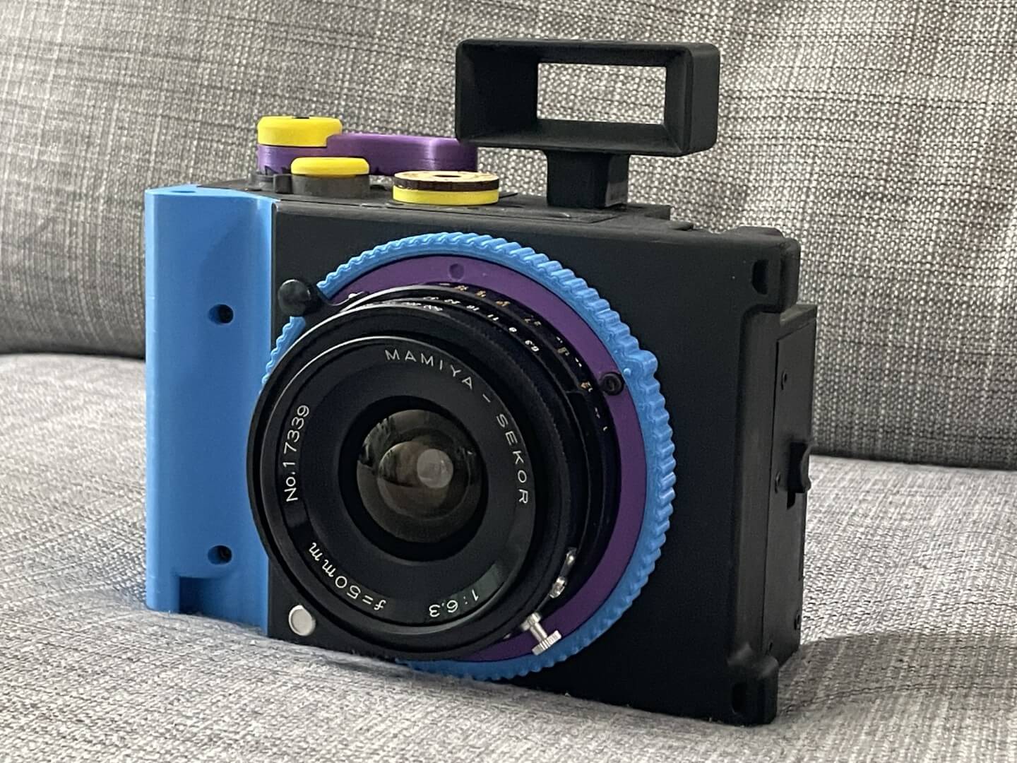 Brancopan camera, with a blocky appearance, printed in purple, yellow, cyan, and black, sitting on a couch with a Mamiya Press 50mm lens attached to it