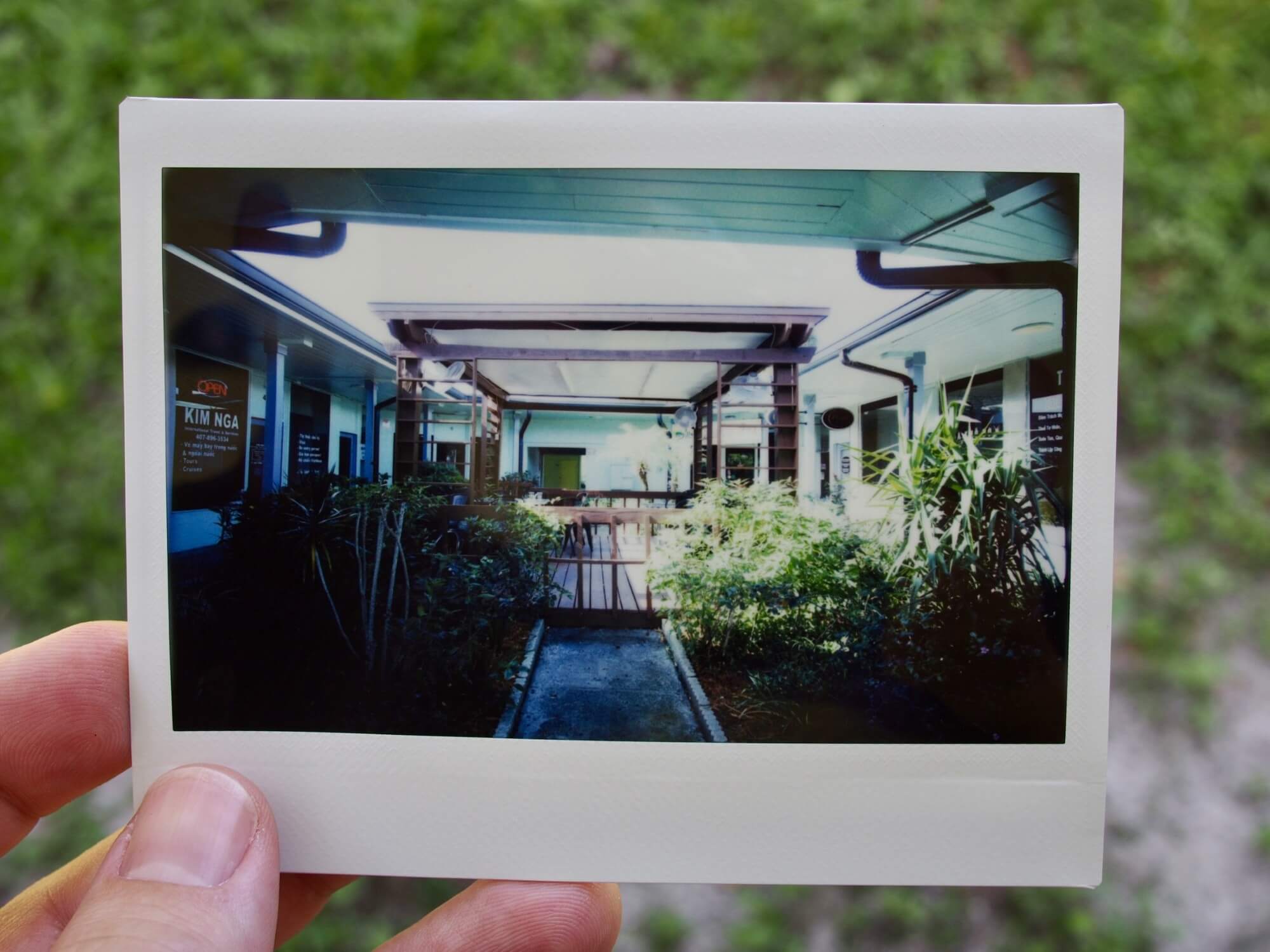 Wide angle Instax Wide print of a courtyard with a wooden gazebo in the middle, surrounded by plants and the entrances to businesses